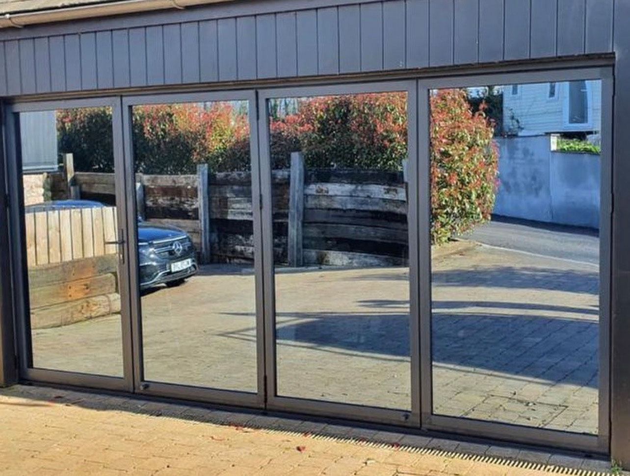Exterior view of closed four-panel bi-fold doors with tinted, reflective glass and dark frames, reflecting the surroundings.
